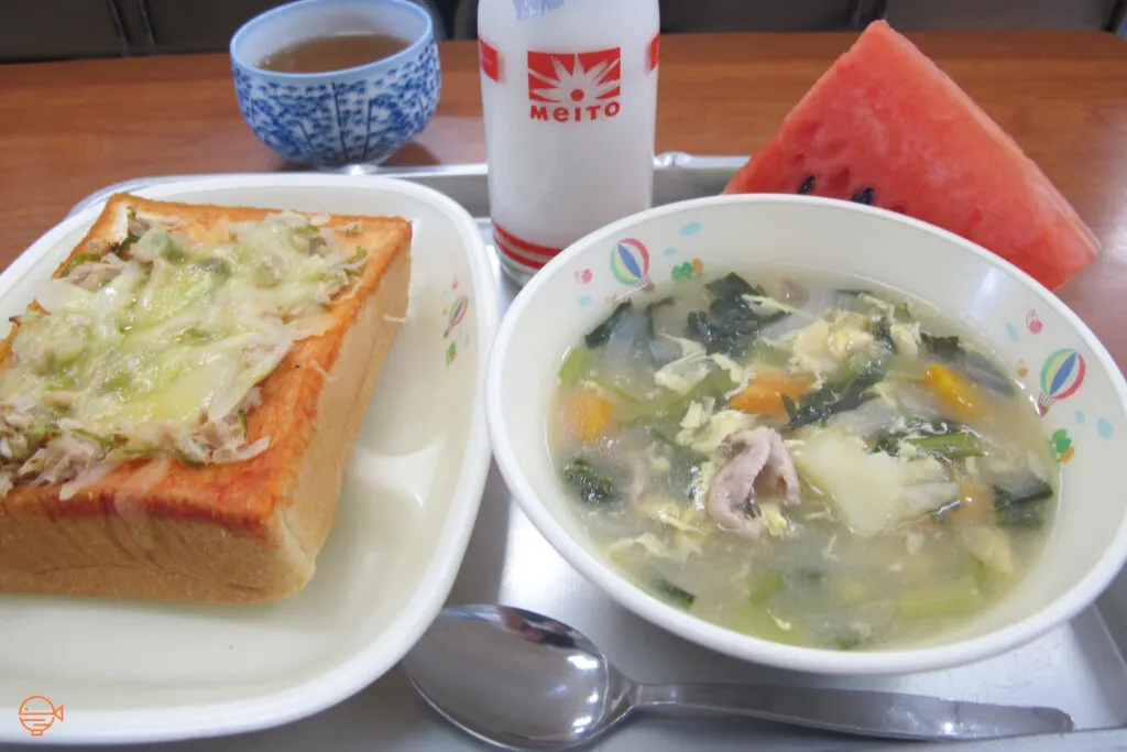 Hot soup with pork, seaweed and vegetables. To the left is a thick slice of bread with a tuna, vegetable and cheese topping. At the back is a big slice of watermelon, a bottle of milk and hot hojicha (Japanese roasted green tea).