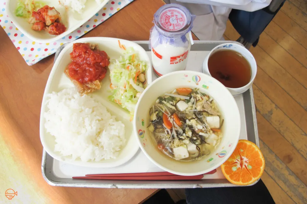 Fried fish with a tomato-based sauce, a cold pickled salad and a serving of rice. To the right is a large bowl of vegetable, tofu and pork soup, along with half an orange, a cup of hot hojicha tea and a bottle of cold milk.