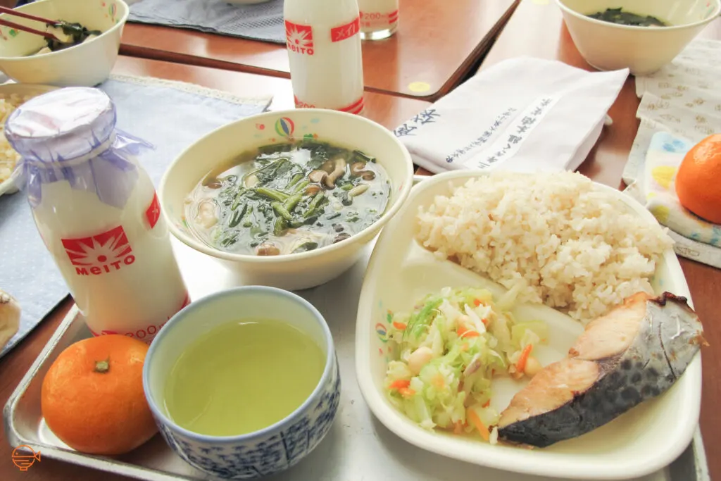 A serving of rice with a piece of salmon and a cold pickled salad. To the left is a hot bowl of soup filled with green vegetables and mushrooms, a cup of green tea, a mandarin and a bottle of cold milk.