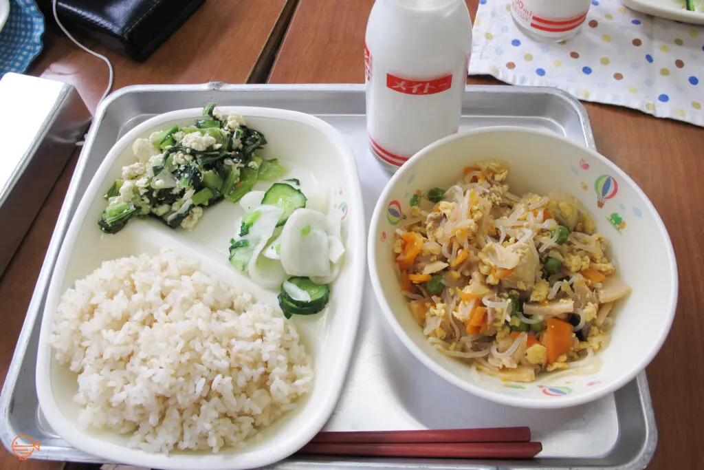 A serving of rice with a green vegetable and tofu salad, and pickled daikon radish and cucumber. To the right is a large bowl of glass noodles with vegetables and egg. Behind it is a bottle of cold milk.