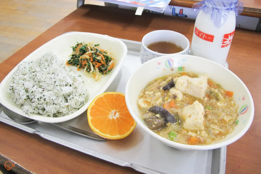 Seasoned rice with a cold pickled salad. To the right is a large hot bowl of soup filled with vegetables, tofu and pork. Sitting around it is half an orange, a hot cup of hojicha tea and a bottle of cold milk.