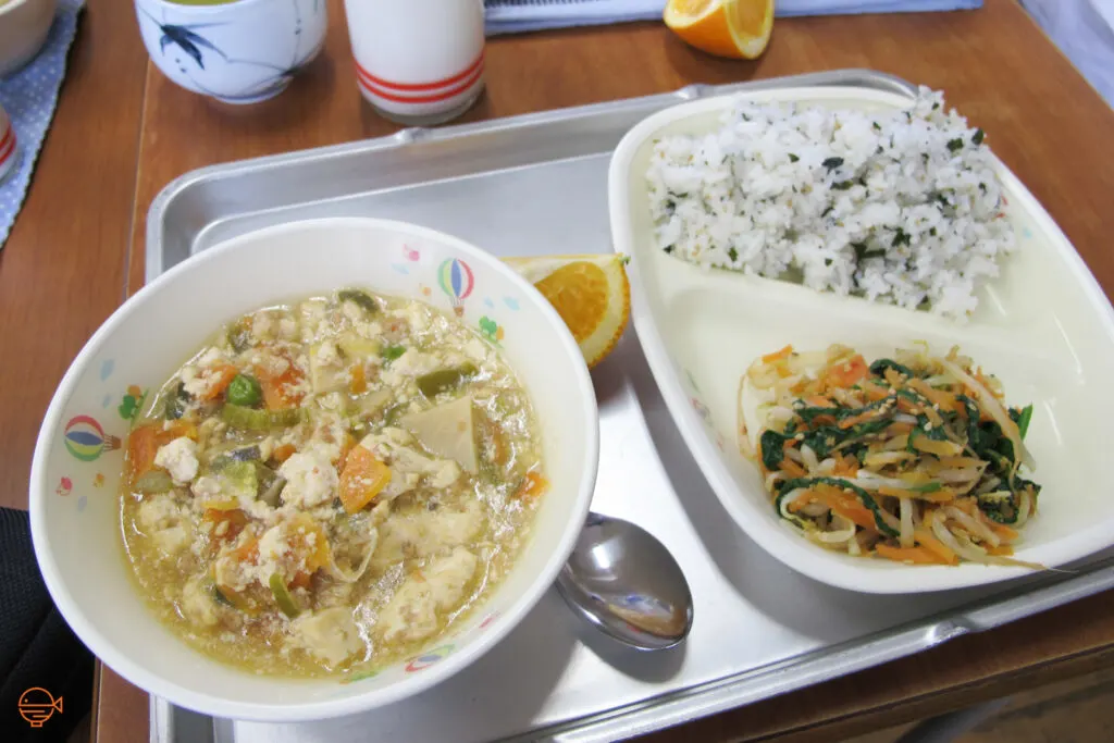 A large bowl of hot soup with tofu and vegetables. To the right is a serving of seasoned rice, a cold pickled salad and a slice of orange, with a hot cup of Japanese tea and cold milk at the back.