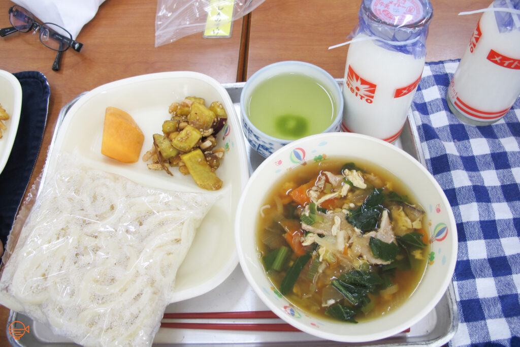 A large bowl of pork, vegetable and seaweed soup with a packet of udon noodles to the left to add to it. Behind that is a sweet potato side salad and a piece of persimmon, along with a hot cup of green tea and a cold bottle of milk.
