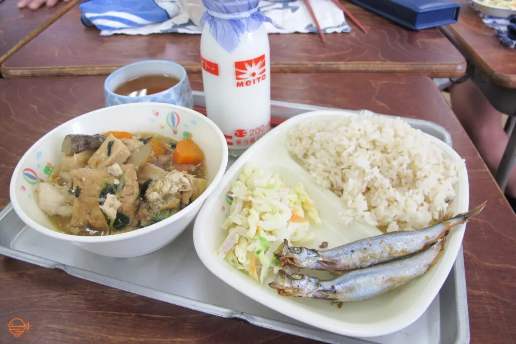 A serving of rice, two grilled smelt fish, a cold pickled salad, a bowl of soup packed with vegetables and tofu, a cup of hojicha tea and a bottle of cold milk.