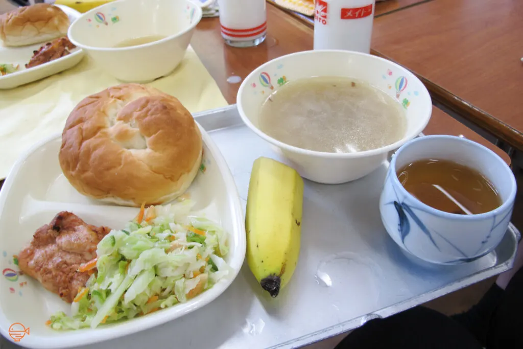 A bread roll, pork and a cold pickled salad, along with a cloudy soup, half a banana, a hot cup of hojicha tea and a glass of cold milk.