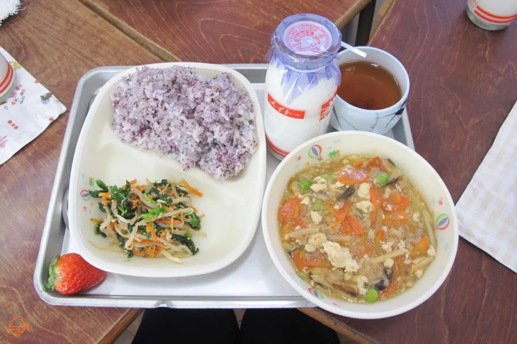 Seasoned rice, a pickled vegetable side salad, a strawberry, a bowl of tofu, pork and vegetable soup, along with a cup of roasted green tea and a bottle of cold milk.