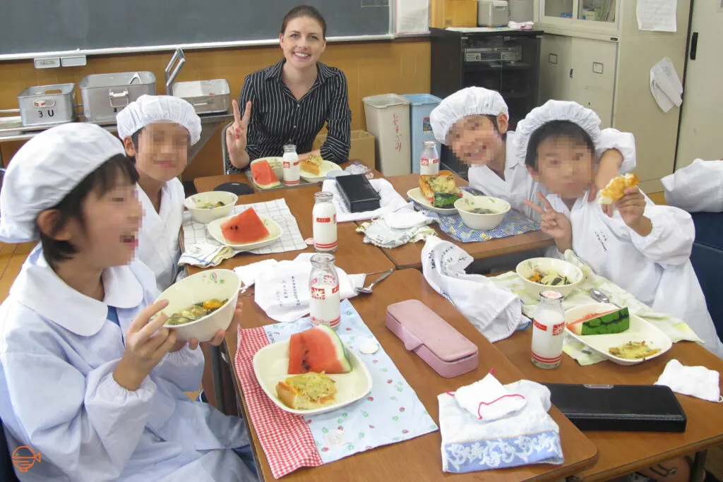 The author sits at a school lunch table with four students in their white school lunch uniforms who are partially through their meals and posing and smiling for the camera.