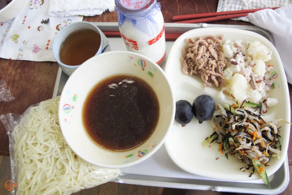 A packet of cold noodles and a soy sauce based dipping sauce, along with a hijiki seaweed and vegetable salad, potato salad with bacon, a serving of pork, two purple grapes, a cup of roasted green tea and a bottle of cold milk.