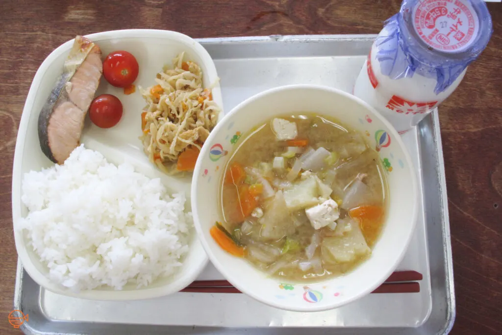 A serving of rice, fish, traditional side salad and two cherry tomatoes, along with a vegetable, tofu and pork soup, and a bottle of milk.