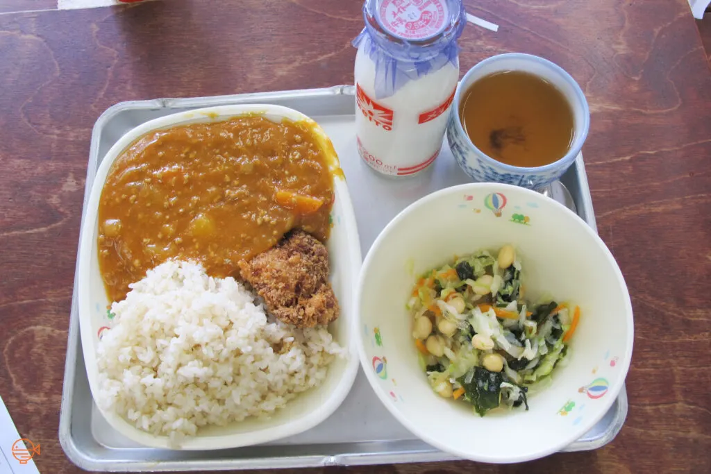 Curry with rice and fried pork, along with a cold pickled salad, a hot cup of roasted green tea and a bottle of cold milk.