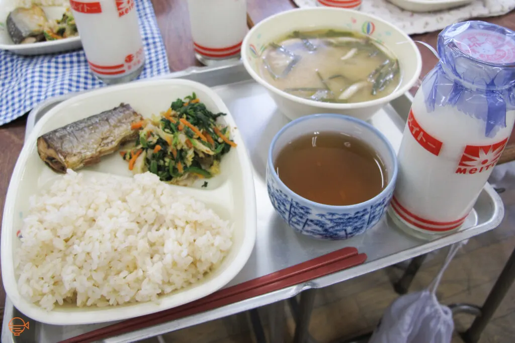 A serving of rice, fish and vegetables, along with a mushroom and seaweed soup, and a cup of roasted green tea and a bottle of milk.