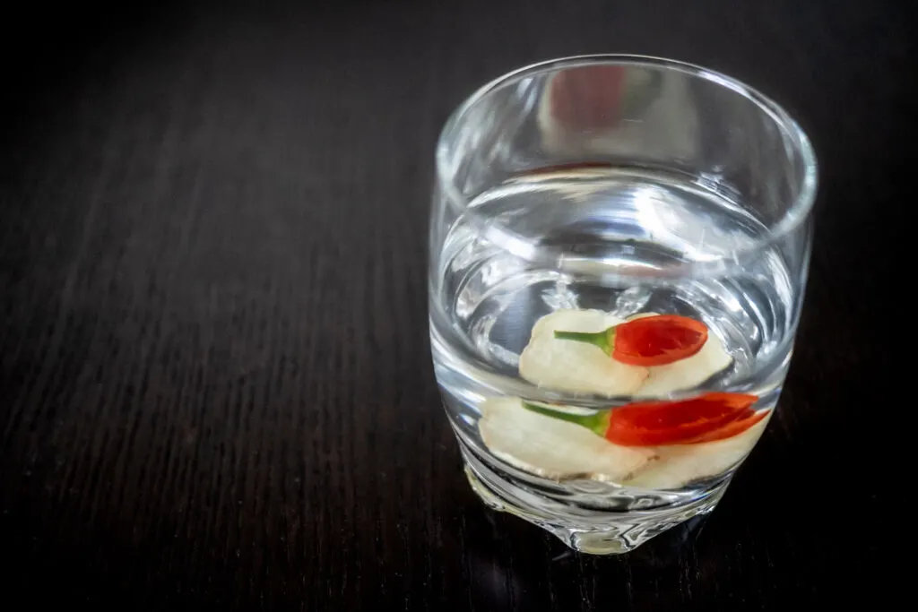 Shochu served oyuwari style with hot water and some sliced ginger and chili in a clear glass on a dark wooden counter top.
