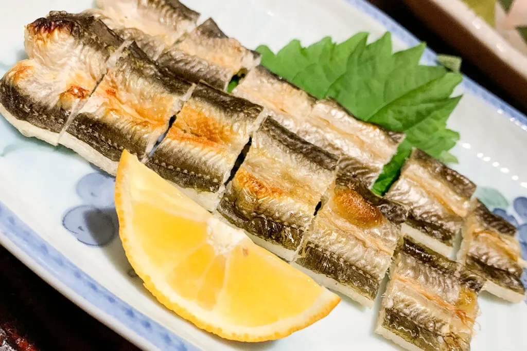 Grilled anago eel on a plate served with a shiso leaf and a wedge of lemon.