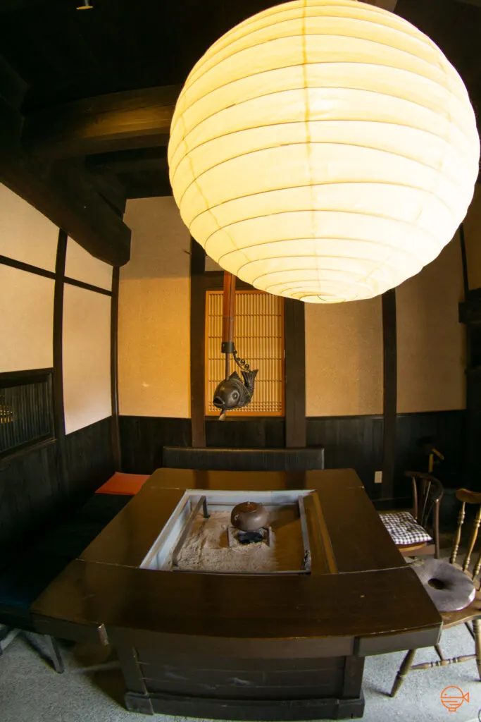 A wooden hibachi table with chairs around it. Set into the middle of the table is a metal box filled with warm ash and in the center is a small pile of burning charcoal with a tea pot on top.