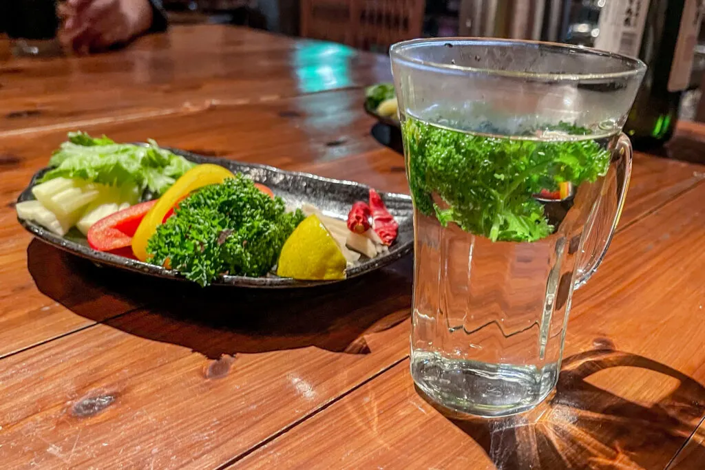 A plate of various infusions to add to a shochu cocktail, including celery, bell peppers, parsley, lemon, ginger and chili, next to a glass of parsley-infused shochu.