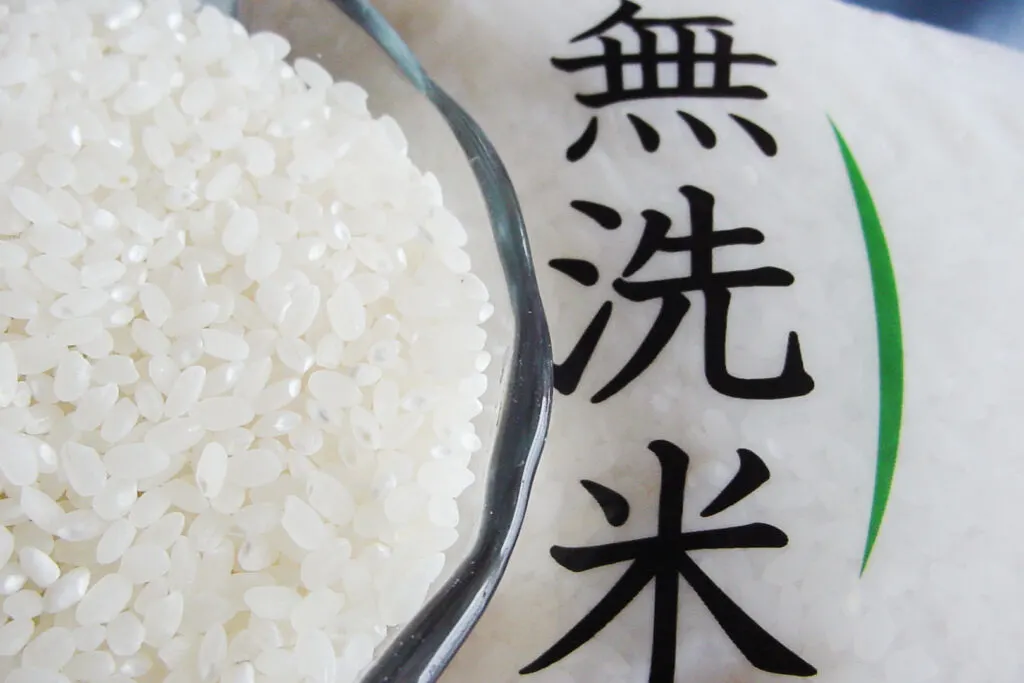 A bowl of uncooked Japanese rice sits on a bag of rice with 'musenmai' (no-wash rice) written on it in kanji characters.