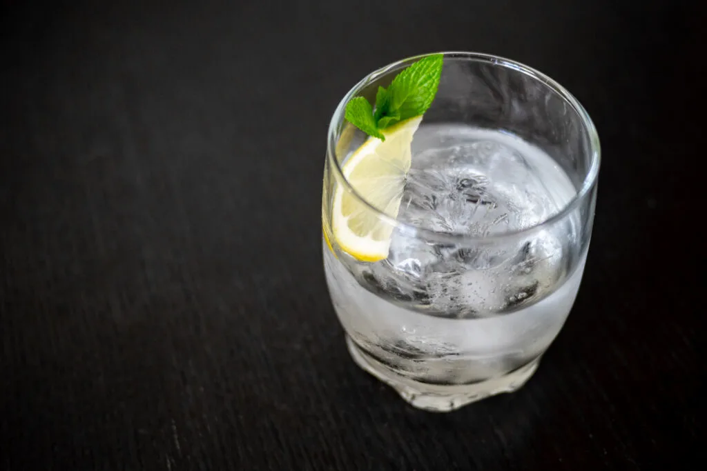Shochu served mizuwari style in a clear glass with ice and water, along with some lemon and mint sits on a dark wooden counter top.