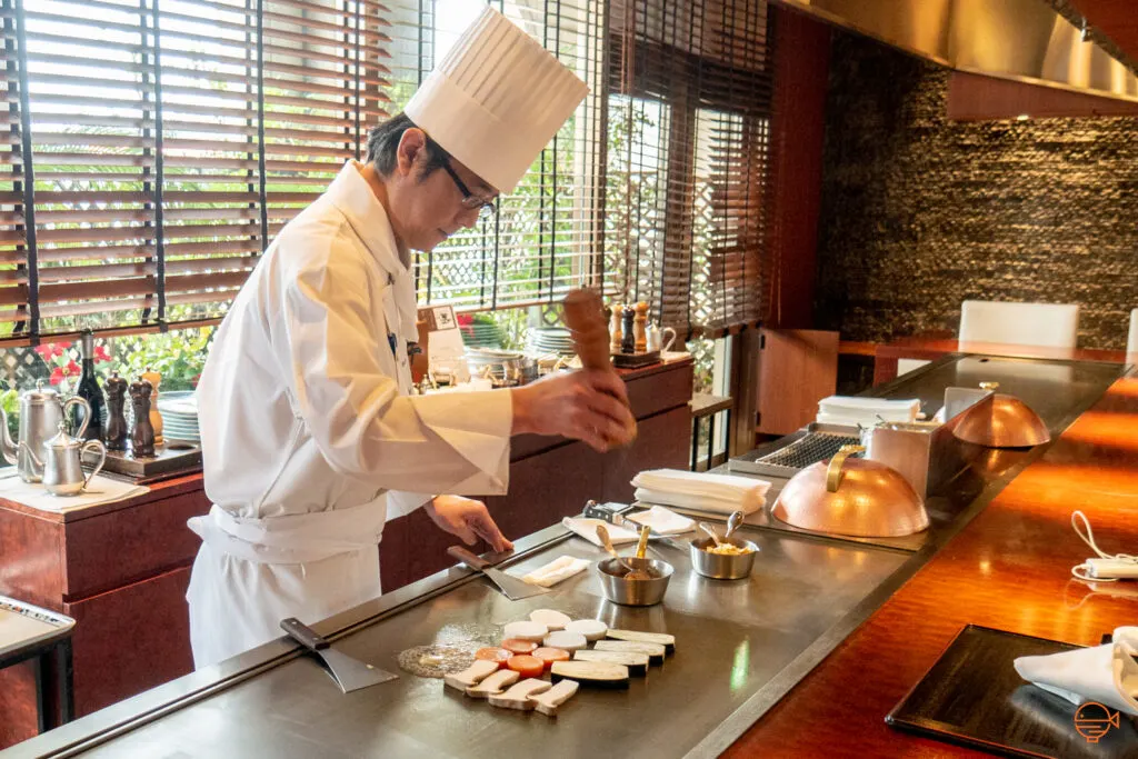 A Japanese chef with a white chef's jacket and tall hat is seasoning teppanyaki vegetables over a Japanese teppan grill.