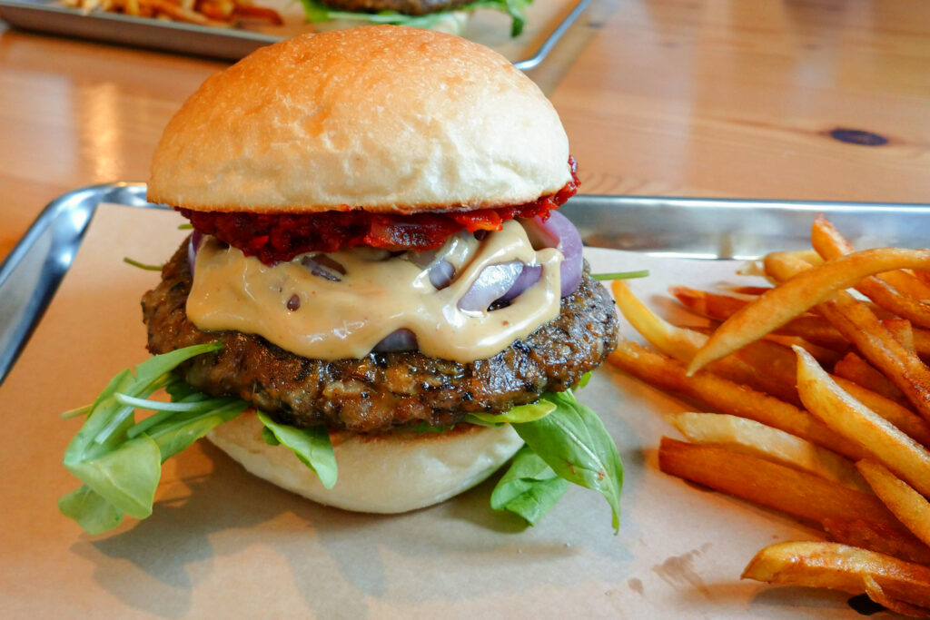 A plump burger with a vegan patty, cheese sauce, tomato jam, marinated onions, baby greens and mayonnaise sits on a tray with a side of fries. Photo by Kaori, S.