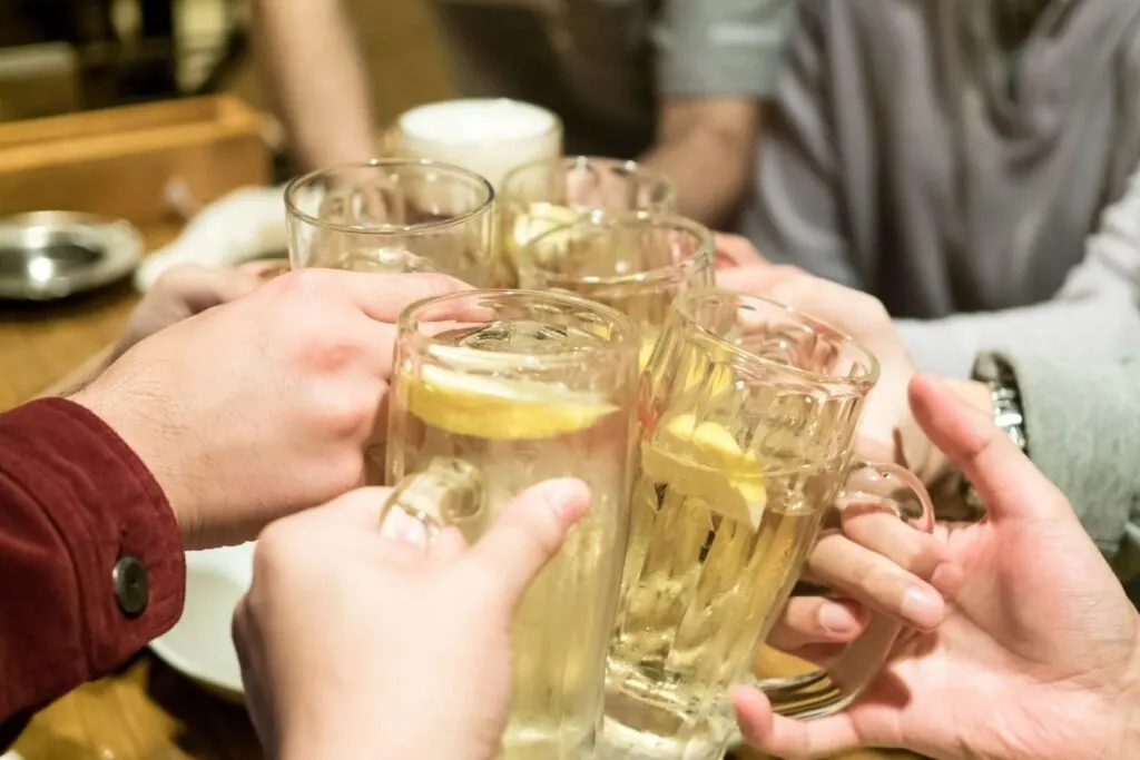 Five people each hold a glass of chu hi with a slice of lemon in it at a bar, clinking their glasses together at the center of the table in a 'cheers' motion.    
