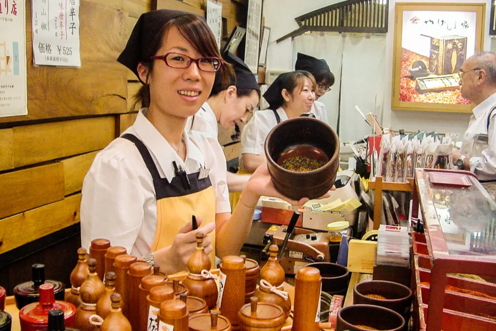A female staff member at Yagenbori Shichimi Togarashi in Asakusa (Tokyo), faces a wooden bowl towards the camera to show the custom blend she is making for the author. Behind her are three other staff members working; one is assisting an older man in the background. In the foreground are variously-shaped wooden containers for storing your 7 spice mix.