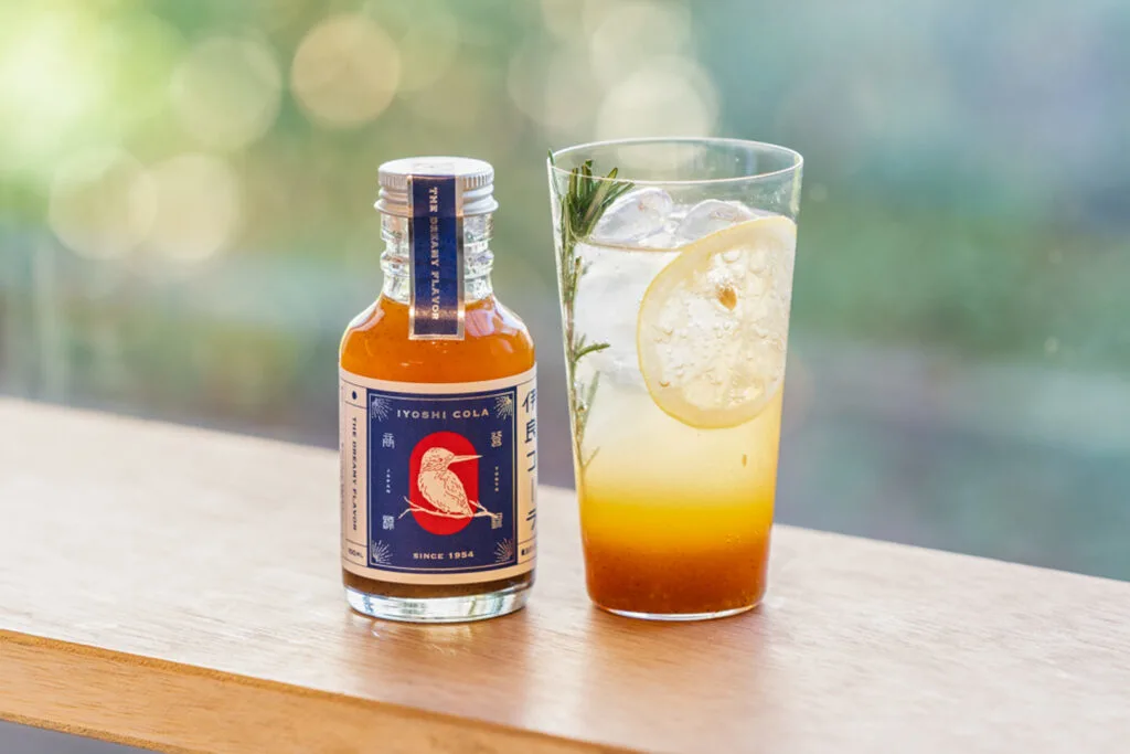 An unopened bottle of Iyoshi Cola with a tall glass to the right showing a serving suggestion: some orange-colored Iyoshi Cola syrup with water, ice, a slice of lemon and a sprig of rosemary.