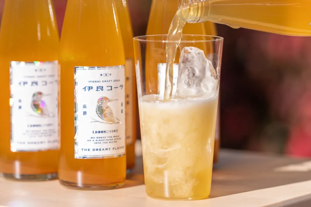 In the background, bottles of Iyoshi's "The Dreamy Flavor" Craft Cola are lined up, while in the foreground a glass is being prepared to drink with the orange liquid being added to a glass of ice and possibly soda water.  