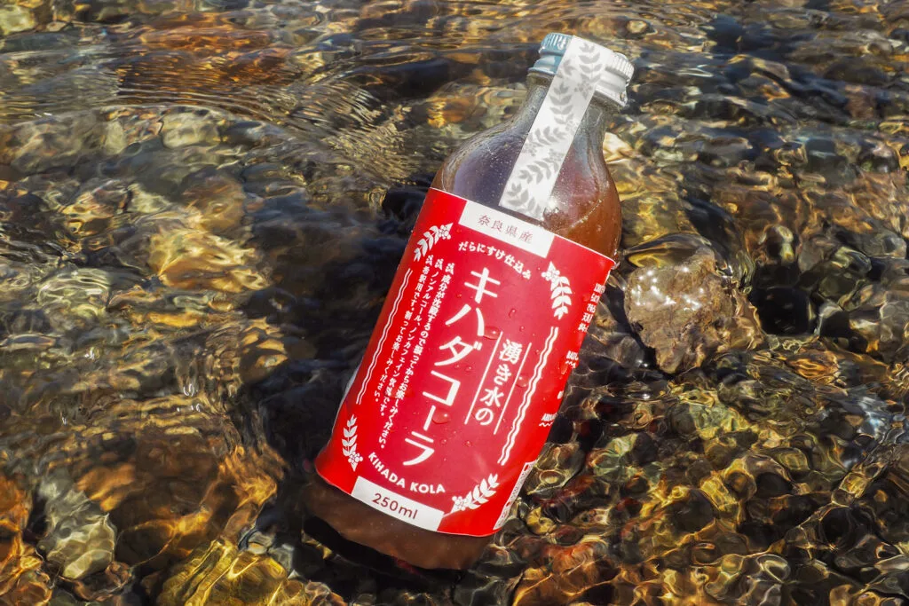 A bottle of brown-colored Kihada Cola concentrate with a red label sits partially in the water and against a rock of a shallow river bed with crystal-clear water.