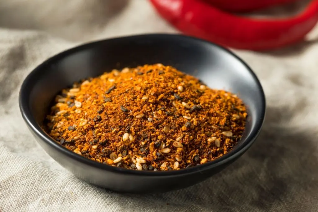 A black ceramic bowl filled with orange-red colored shichimi togarashi (Japanese 7 spice mix), with a hot red capsicum (Capsicum annum) behind it.