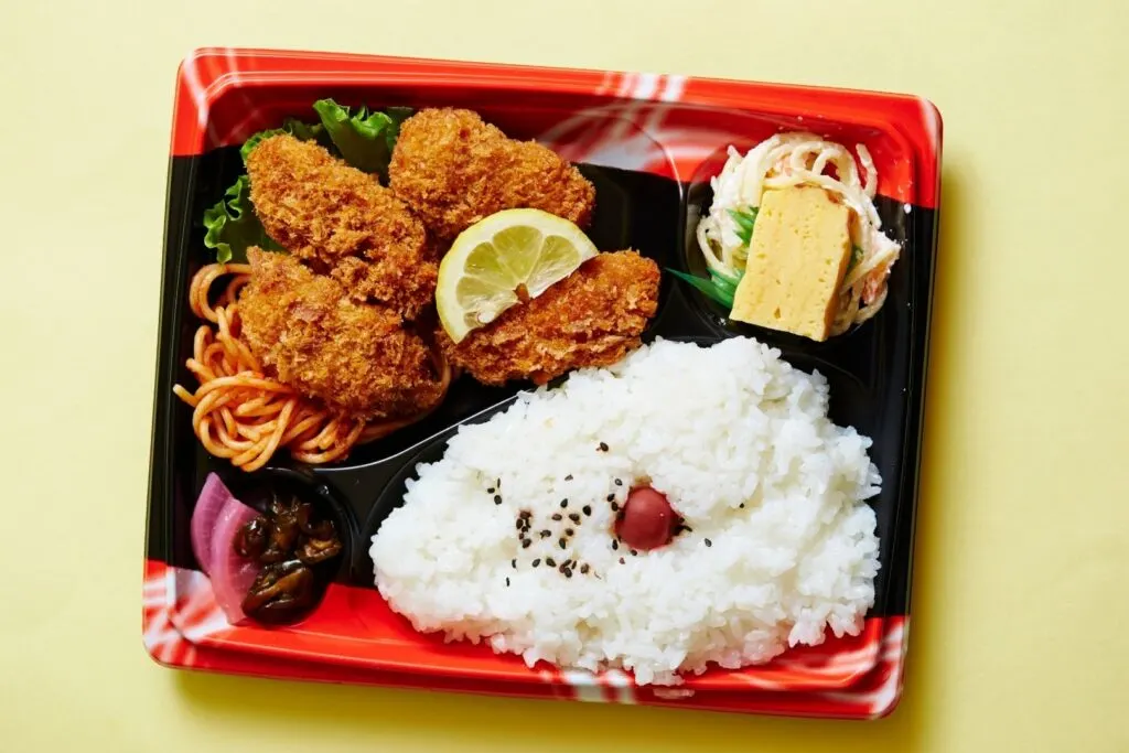 A classic Japanese store-bought bento box featuring fried oysters. The egg and the saucy salad are separated by plastic sushi grass.