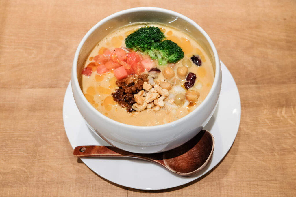 A bowl of Ultimate Vegan Ramen at Tokyo Noodle Stand in Harajuku. It has a creamy golden broth with broccoli, beans, tomato and even peanuts, all in a very contemporary rounded white bowl with a brown wooden spoon. 