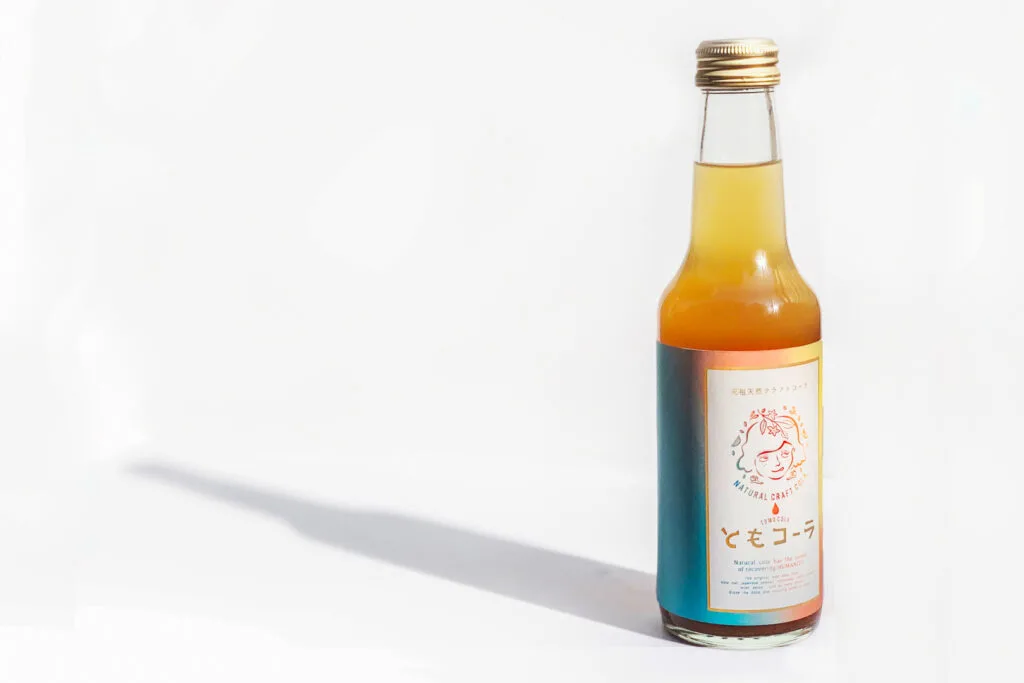 A tall, sleek bottle of orange-colored Tomo Cola is to the right of frame and casting a long shadow against the white surface it's sitting on.