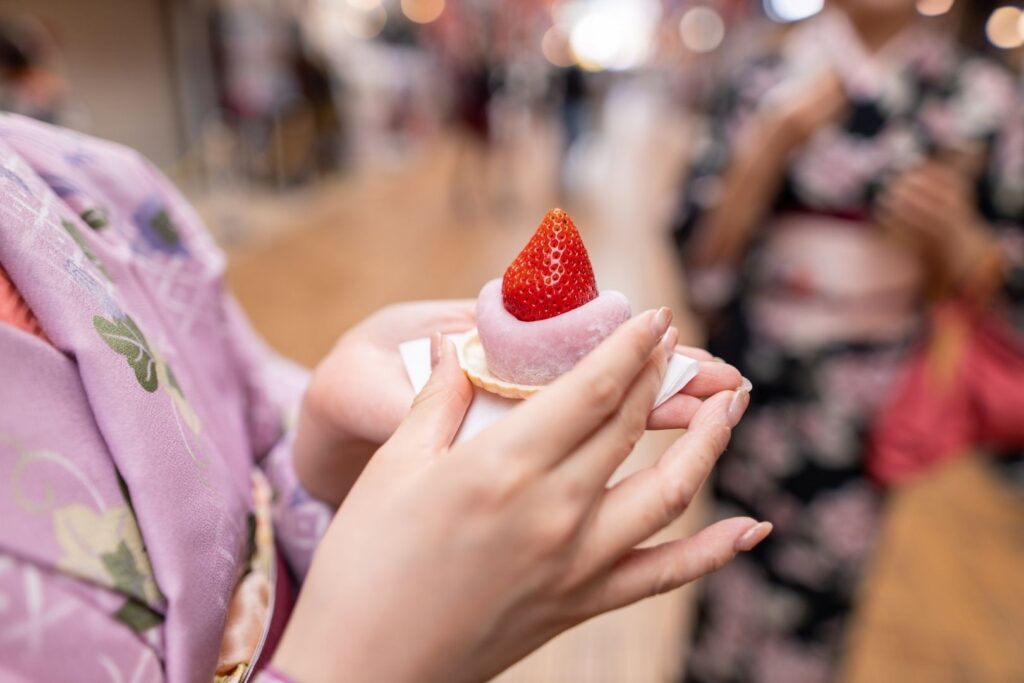 A woman in a lilac-colored kimono holds a pink strawberry daifuku mochi daintily in her hands. Another woman in a pink and black kimono can be seen in the background.