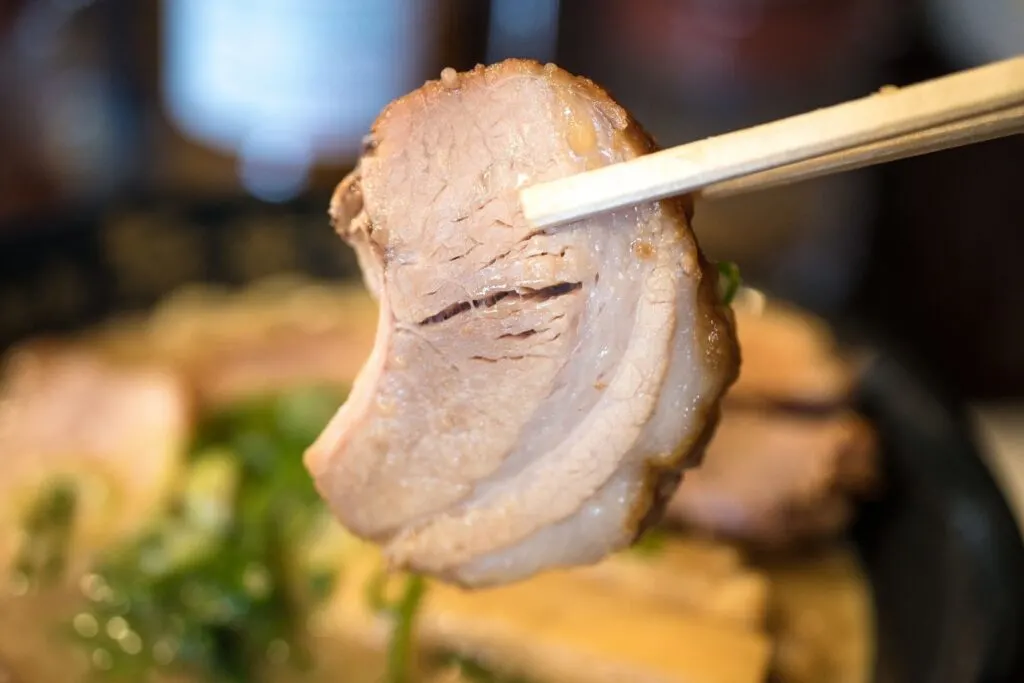 A bowl of ramen is in the background, while a slice of chashu (braised pork) is being held up to the camera with wooden chopsticks from the right.