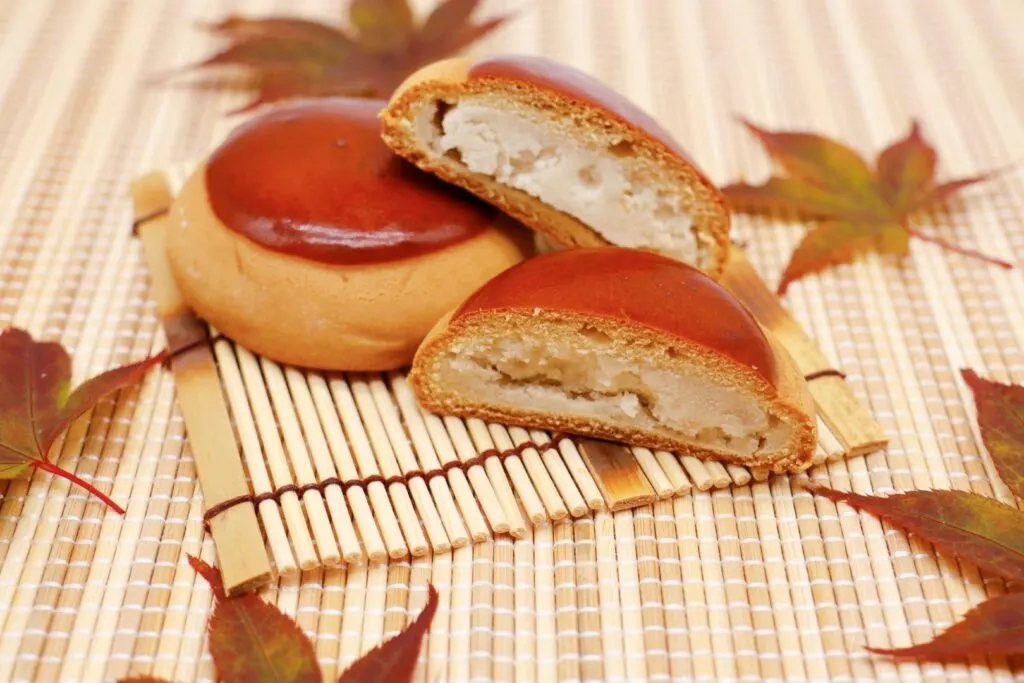 Two chestnut manju on a bamboo place mat surrounded by autumn maple leaves. One of the manju is cut in half showing the chestnut filling.