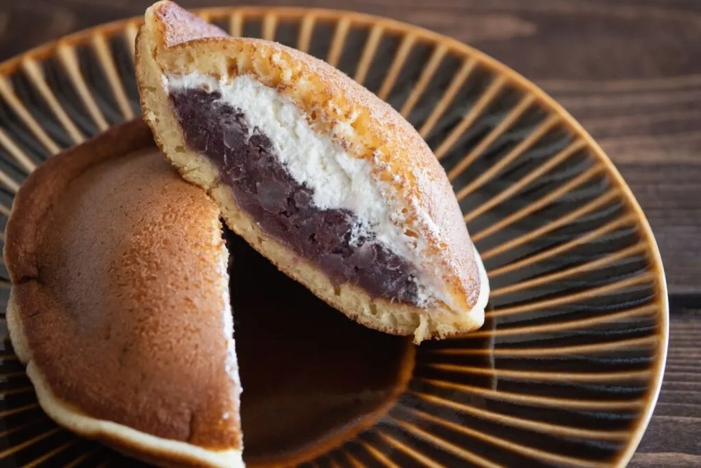 Dorayaki (two small pancakes sandwiched together) with an adzuki bean paste and cream filling cut in half to show the filling and sitting on a brown plate.