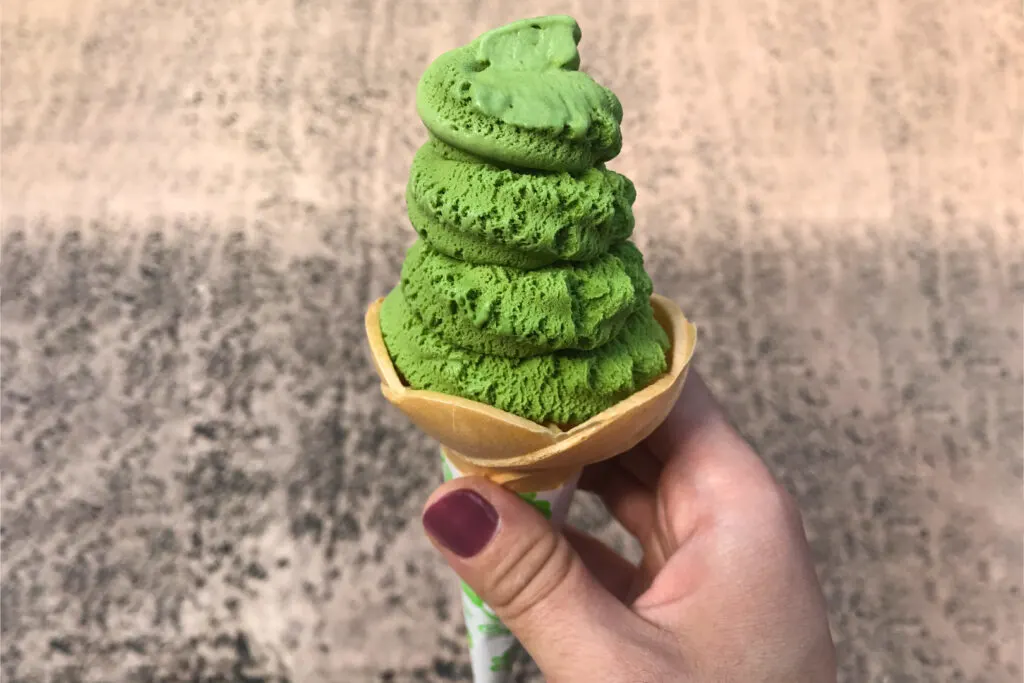 A hand with burgundy nails holds a matcha ice cream cone with a bite already taken from the tip.