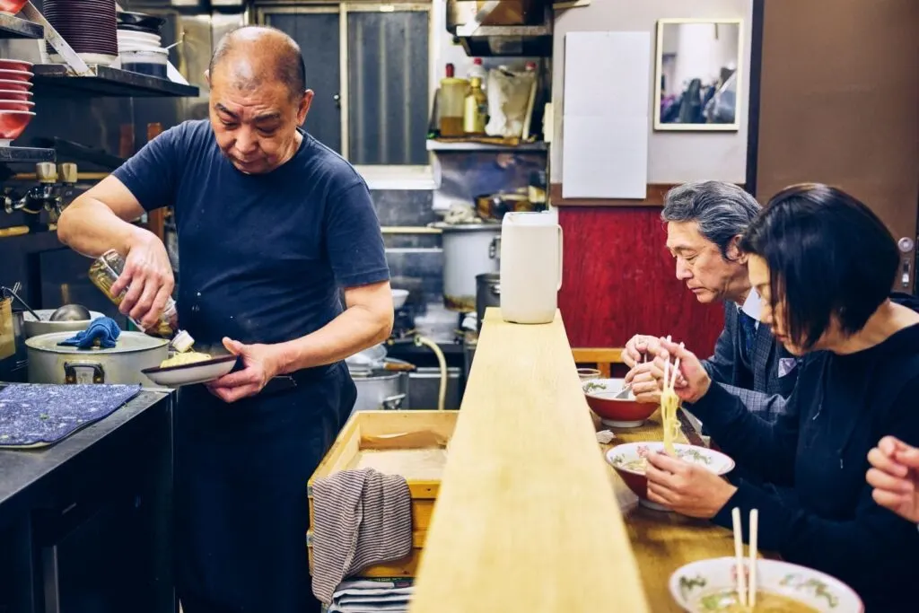 A small ramen shop with the proprietor preparing noodles on one side of the counter while three customers tuck into their ramen bowls on the other.