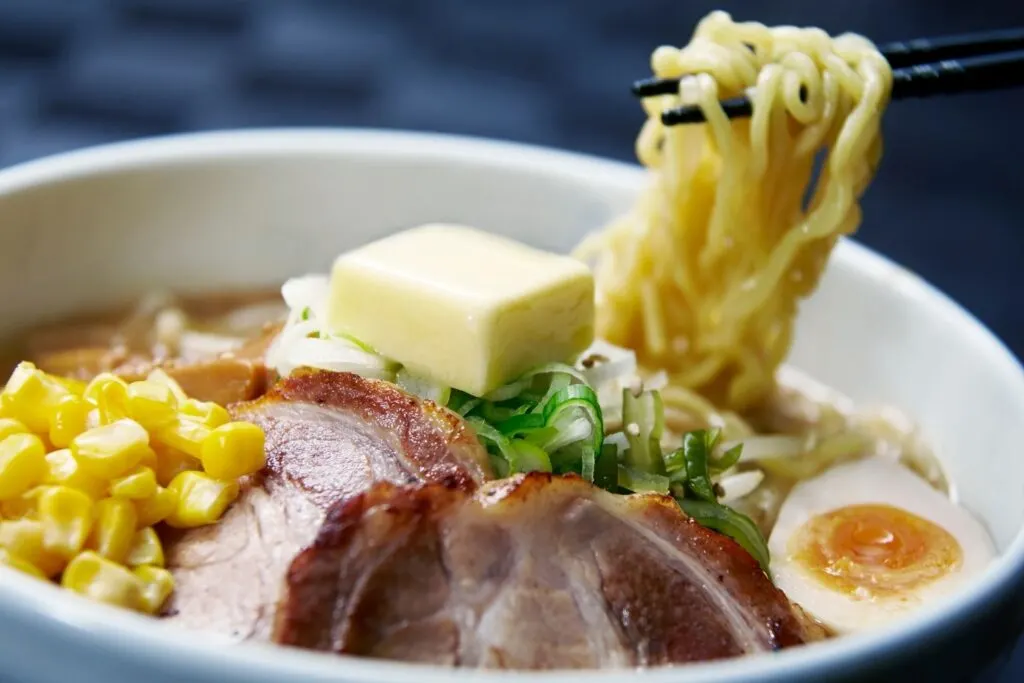 A bowl of miso ramen in Sapporo with corn, charshu sliced pork, egg, spring onions and a square piece of butter on top. Chopsticks are holding up noodles from the bowl from the right.