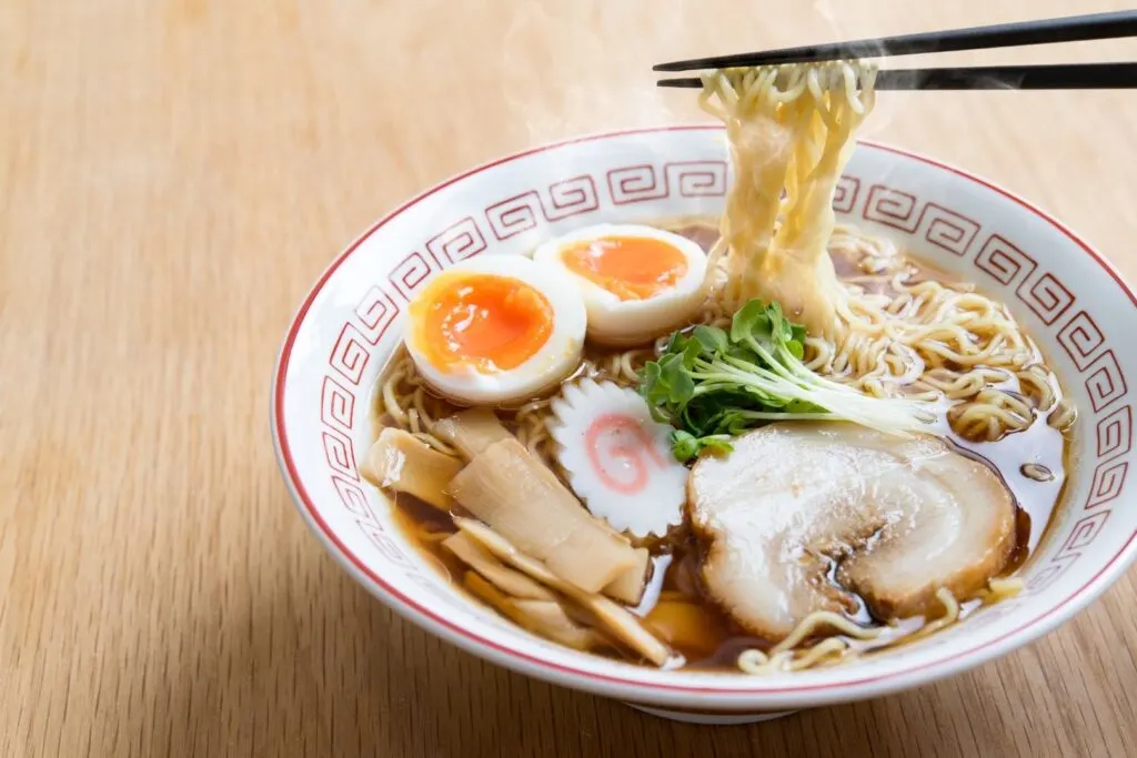 A bowl of shoyu ramen (soy sauce base) with egg, sliced pork, fermented bamboo shoots, narutomaki and some greens as toppings. Chopsticks can be seen holding up some noodles from the right.