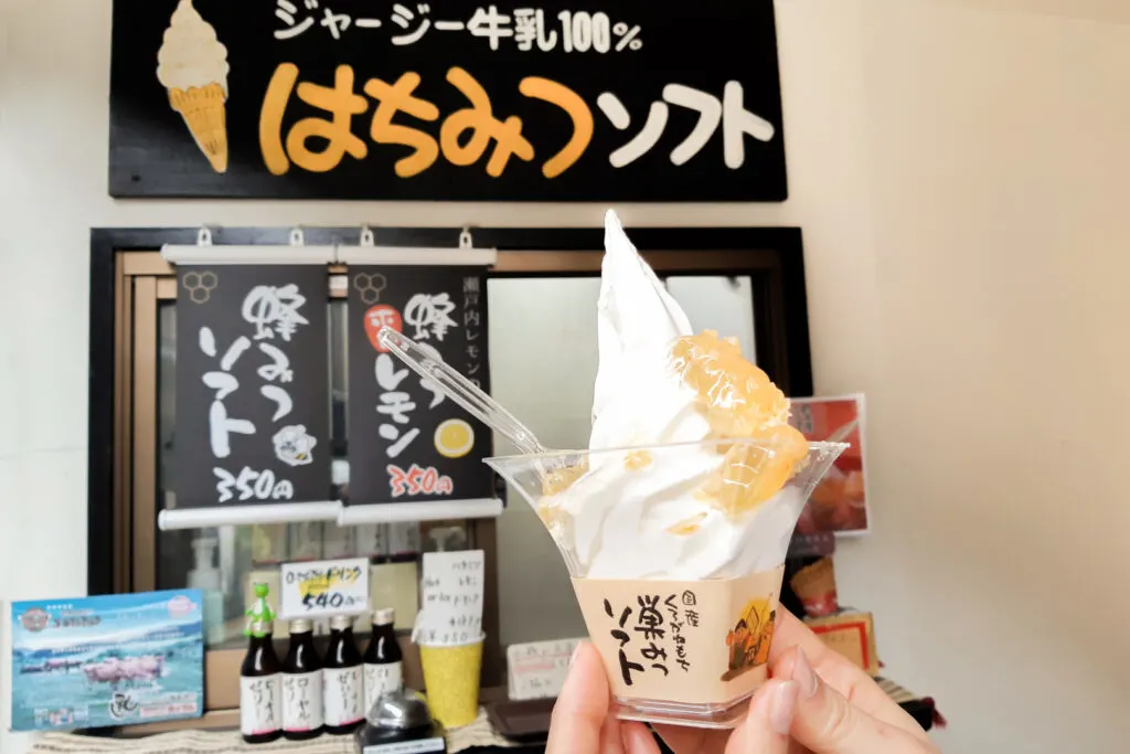Two hands can be seen holding a soft-serve ice cream in a cup made with 100% jersey milk and topped with Japanese honey still in the honeycomb in front of signs for the product at Hana-no-Michi, Shuzenji.