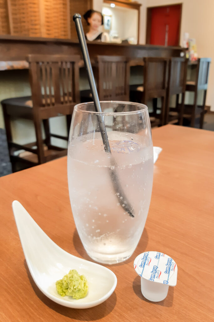 A glass of soda water with a long, black spoon standing up in it. In front of the glass is a Chinese-style spoon with freshly-grated wasabi on it and next to it is a plastic, disposable container of gum syrup with a peelable lid.  