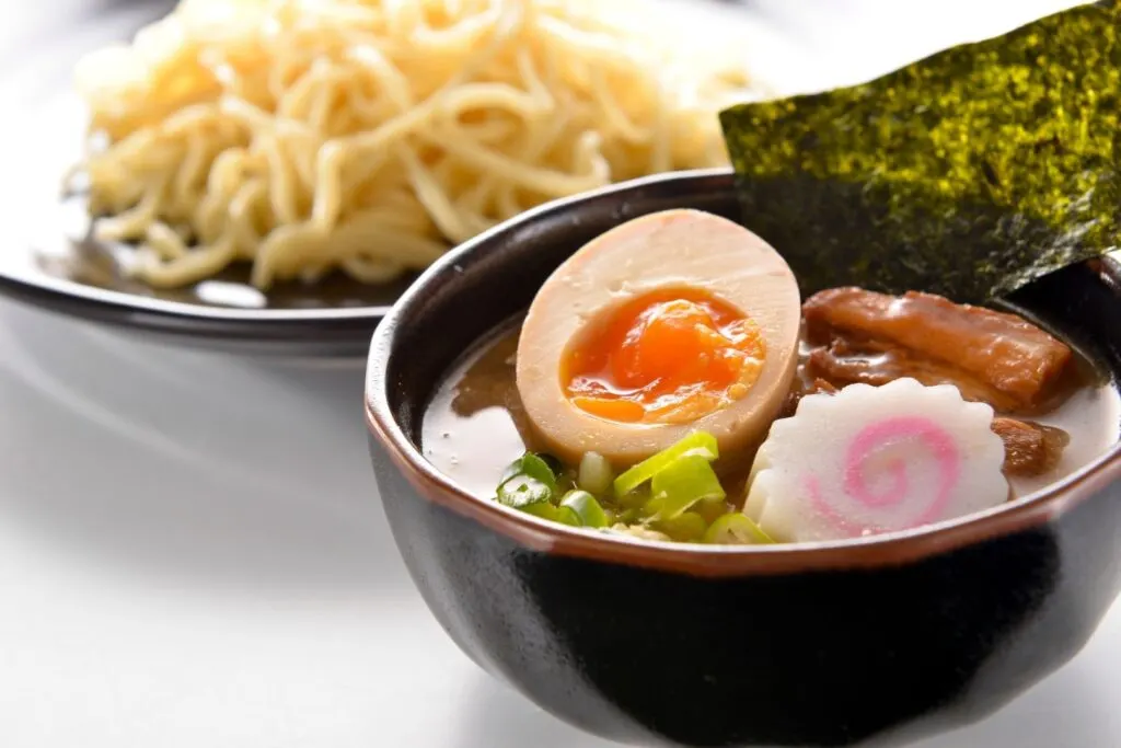 Tsukemen - a type of ramen where the noodles are served separately to the broth. A plate of ramen noodles can be seen in the background, while a bowl of broth with various toppings including a piece of nori (dried seaweed) sticking up at the side, can be seen in the foreground.