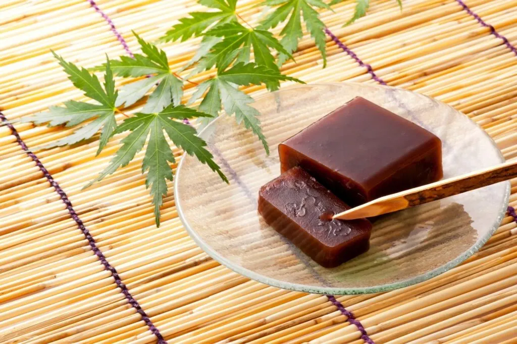 A dark-brown rectangular slab of yokan with one slice cut off the end and resting on the clear plate beneath it. The plate is sitting atop a bamboo placemat and green momiji leaves are sitting behind it as seasonal decoration.