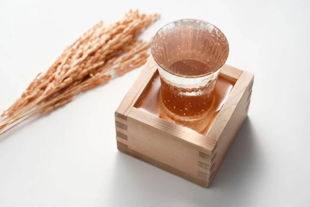 Sake overpour: A sake glass sits inside a masu. Both the glass and the wooden masu box are filled to capacity, the sake having been poured until it overflows into the masu. Ears of rice are on the counter top to the left of the sake.