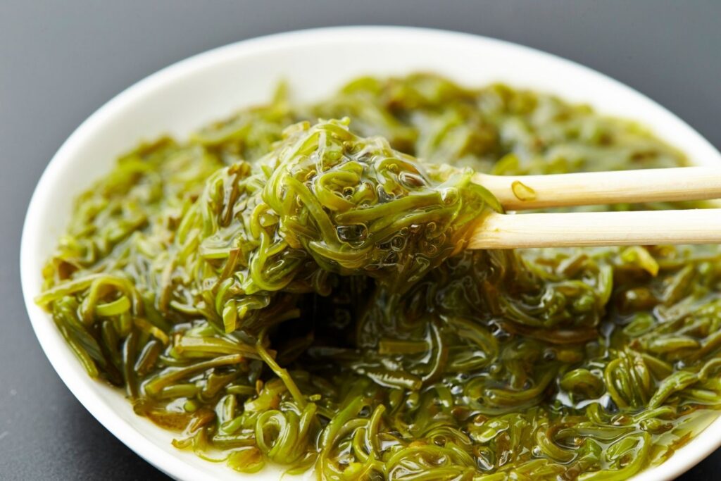 A white bowl of thin, green-colored mekabu on a gray surface. Chopsticks hold up some for the camera from the right.