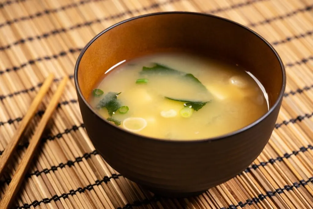 A bowl of miso soup with seaweed, tofu and spring onions in a wooden bowl on a bamboo place mat with a pair of wooden chopsticks to the left.