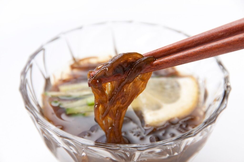 A glass bowl on a white surface half-filled with mozuku Japanese seaweed in vinegar. A thin slice of lemon and some greens can also be seen in the bowl. Chopsticks hold up some of the thin, brown seaweed for the camera from the right.