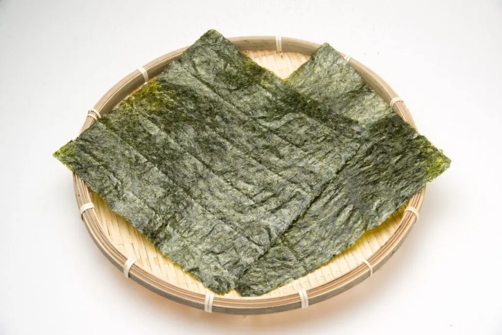 Two large sheets of dried nori Japanese seaweed sit in a shallow bamboo basket on a white surface.