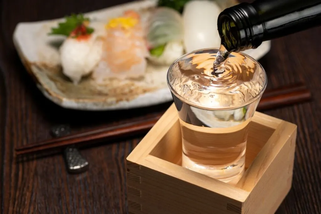 Sake in a box: A glass is set inside a wooden masu cup and is being filled to overflow. Behind it is a plate of sushi.